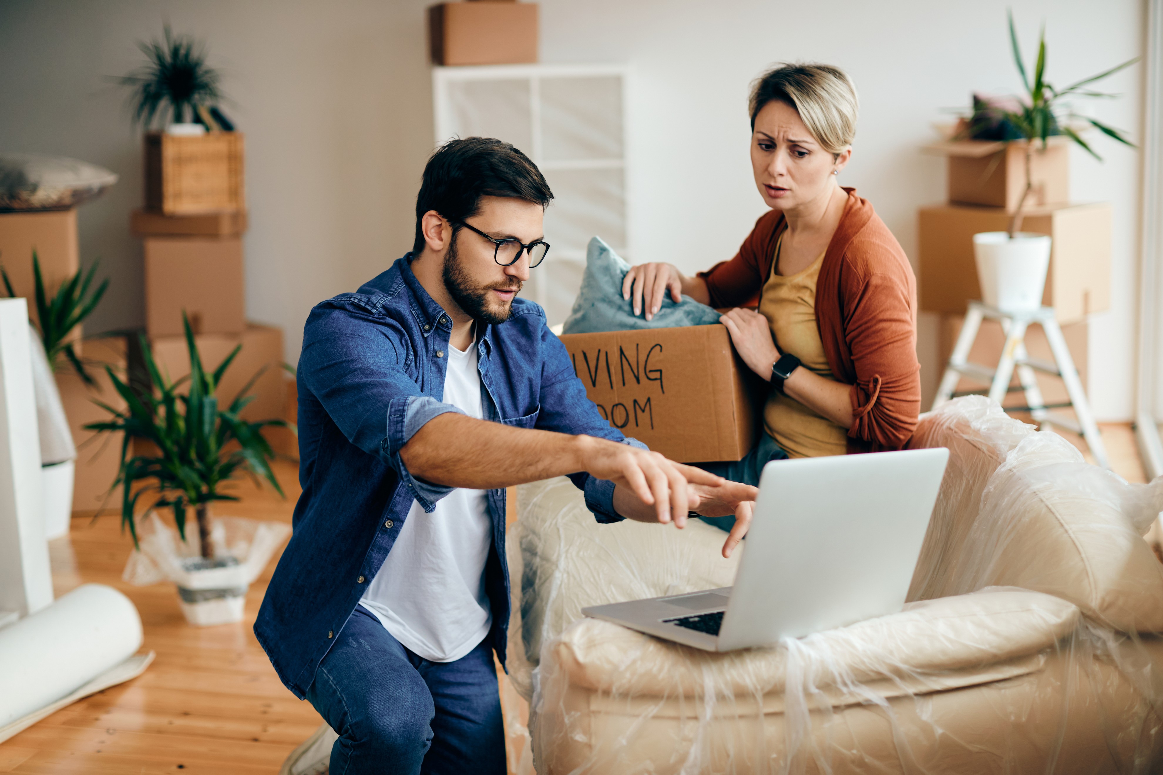 Concerned couple researching home-buying pitfalls on a laptop amidst moving boxes.